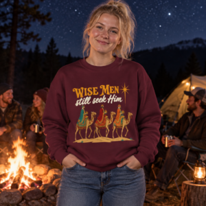 Woman wearing a maroon Wise Men Still Seek Him Gildan 18000 Christmas sweatshirt beside a campfire